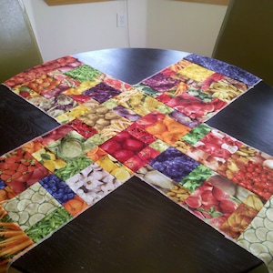 May include: A colorful table runner with a patchwork design featuring various fruits and vegetables. The runner is made of fabric squares sewn together in a cross pattern. The runner is laid out on a black table.