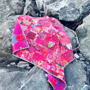 Op de afbeelding: Een levendige patchworkquilt met een verscheidenheid aan roze, rode en magenta stoffen. De quilt heeft bloemen- en geometrische patronen, met een zwart-wit gestreepte rand. De quilt is over grijze rotsen gedrapeerd.