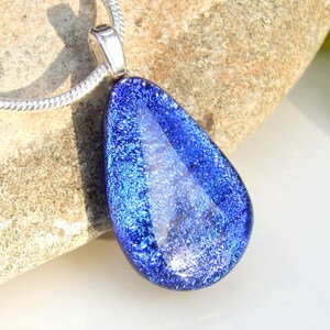 Puede incluir: Un collar con un colgante en forma de l&aacute;grima de vidrio azul. El vidrio tiene un acabado brillante e iridiscente.
