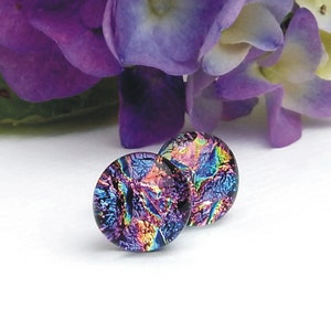 May include: A pair of round dichroic glass stud earrings. The earrings have a dark purple base colour with iridescent blue, green, and gold accents. The earrings are displayed on a white surface with purple flowers in the background.
