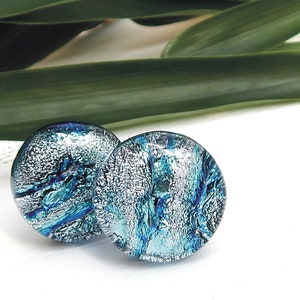 May include: A pair of round dichroic glass stud earrings. The earrings feature a mix of silver and shades of blue, creating a shimmering, textured effect. The earrings are set against a white background with green leaves.