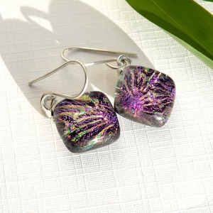 May include: A pair of silver dangle earrings with square-shaped purple and green dichroic glass pendants.
