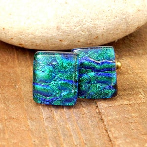 May include: A pair of square-shaped glass earrings with a blue and green iridescent finish. The earrings have a gold-toned metal backing.