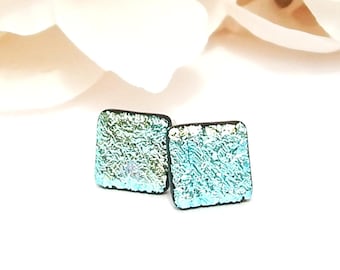 Peut inclure: Une paire de boucles d'oreilles carrées avec une surface texturée et irisée. Les boucles d'oreilles ont un bord noir et une couleur aqua et verte chatoyante. Le fond est blanc avec un élément beige flou.