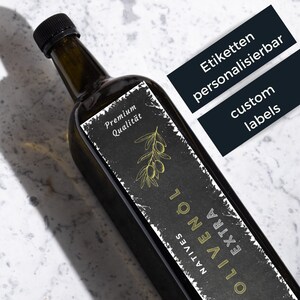 Op de afbeelding: Een donkergroene glazen fles olijfolie met een zwart-wit etiket waarop "Natives Olivenöl Extra" en "Premium Qualität" staat. Het etiket bevat ook een tekening van een olijftak met olijven. De fles staat op een wit marmeren oppervlak.