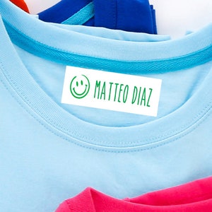 Può includere: Una maglietta blu chiaro con un bordo turchese intorno al collo. Un'etichetta bianca termoadesiva con una faccina sorridente verde e il testo "MATTEO DIAZ" è attaccata al collo.