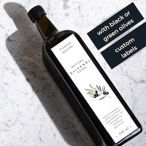 Puede incluir: Una botella de vidrio de aceite de oliva con una etiqueta blanca que dice "Natives Olivenöl Extra" y "Premium Qualität". La etiqueta también presenta un dibujo de una rama de olivo con aceitunas negras. La botella está sobre una superficie de mármol blanco. El texto "with black or green olives" y "custom labels" está escrito en negro sobre rectángulos blancos.