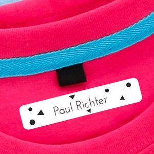 May include: White rectangular label with black text and symbols. The label reads "Paul Richter" and has two black dots, a triangle, and a small arrow on each side. The label is attached to a pink shirt with a blue trim.
