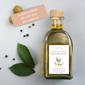 Op de afbeelding: Een doorzichtige glazen fles met biologische extra vierge olijfolie met een kurkstop en een wit etiket met de tekst "Superior Quality Organic Extra Virgin Olive Oil" en "Koudgeperst, ongefilterd Biologisch geteeld en handgeplukt".