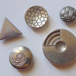 May include: Five silver metal buttons with different designs. The buttons are round, triangular, and have a variety of textures.