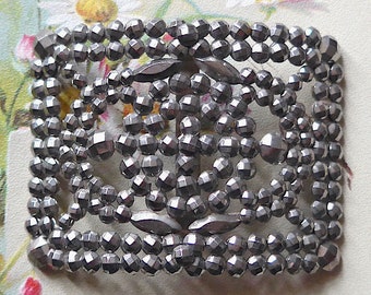 May include: A silver metal buckle with a geometric design made of small, faceted beads. The buckle is rectangular in shape and has a smooth, polished finish.