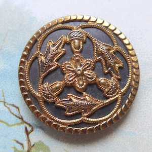 Op de afbeelding: Een ronde, antieke knoop met een goudkleurig metalen ontwerp op een donkergrijze achtergrond. Het ontwerp toont een centrale bloem, bladeren en eikels, omgeven door een touwachtige rand. Deze vintage knoop is waarschijnlijk uit de vroege 20e eeuw.