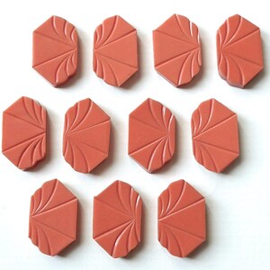 Könnte beinhalten: Zehn korallenfarbene, geometrisch geformte Dekorfliesen. Jede Fliese hat ein einzigartiges Design mit einem fächerartigen Muster auf einer Seite. Die Fliesen sind auf einer weißen Oberfläche angeordnet.