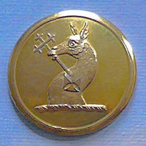 Op de afbeelding: Een gouden metalen knoop met een reliëf van een gestileerd dier met een kruis. Het dier staat op een touwachtig ontwerp.