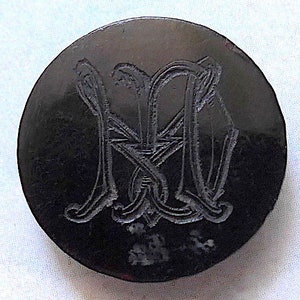 Peut inclure: Un bouton circulaire noir avec un motif gravé de deux 'M' entrelacés au centre.