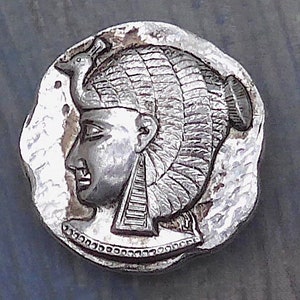 Op de afbeelding: Een zilverkleurige munt met een profiel van een persoon die een uitgebreide hoofdtooi draagt met een vogelmotief. De munt heeft een gedetailleerd reliëfontwerp en een onregelmatige vorm, mogelijk een antiek of een replica. Diameter: ongeveer 2,5 cm.