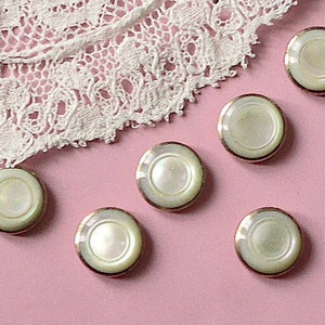May include: Six white pearl buttons with gold trim, arranged on a pink surface.