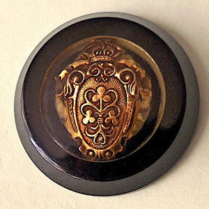 Bottone in lucite, metallo e resina, vintage. Resina nera con uno scudo araldico sotto la lucite, lati inclinati, gambo ad anello. Anni '40 circa.