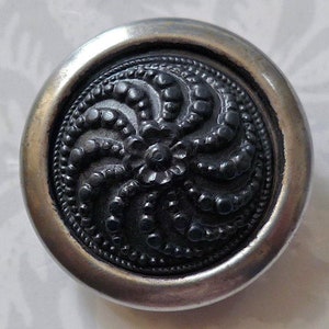 Pode incluir: Um botão prateado com um design circular preto em relevo. O design apresenta um padrão de pontos e linhas em espiral.