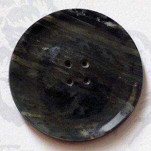 Puede incluir: Un botón redondo de color negro y verde con cuatro agujeros. El botón tiene un patrón de mármol.