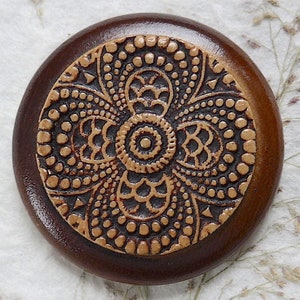 Può includere: Un bottone rotondo in legno con sfondo marrone e un design a mandala beige. Il mandala presenta intricati motivi di cerchi, punti e spirali.