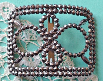 May include: A silver-toned metal buckle with a geometric design made of small, faceted beads. The buckle is designed to be used as a closure for a garment or accessory.