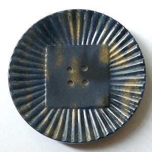Könnte beinhalten: Ein großer, runder Knopf in Blau und Gold mit einem Vierloch-Design. Der Knopf hat eine strukturierte, gerippte Oberfläche.