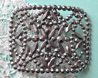 May include: A silver metal buckle with a decorative design of small, round beads and leaf shapes.