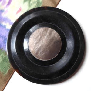 Puede incluir: Un objeto circular negro con un centro plateado. El objeto tiene un diseño estriado y parece estar hecho de un material duro. El fondo incluye un estampado floral en tonos morado, verde y marrón.