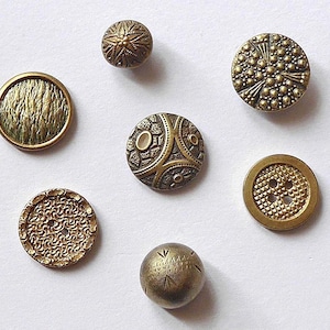 Könnte beinhalten: Eine Auswahl antiker Messingknöpfe in verschiedenen Formen und Texturen. Die Knöpfe weisen runde, gewölbte und kugelförmige Designs mit komplizierten Mustern auf, darunter strukturierte Oberflächen, geprägte Details und geometrische Formen. Die Knöpfe sind auf weißem Hintergrund angeordnet.