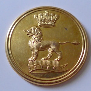May include: A gold-coloured metal button featuring a raised design of a lion standing on a base with a banner and a crown above the lion.