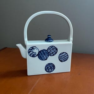 May include: A white ceramic teapot with a rectangular body and a curved handle. The teapot features blue circular designs with various patterns. A small blue lid sits on top. The spout is on the left side.
