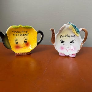 May include: Two ceramic tea bag holders. One is yellow with a black handle and spout, and the text "I WILL HOLD THE TEA BAG". The other is white with a brown handle and spout, and the text "I hold the BAG". Both have smiling faces.