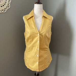 May include: A sleeveless, button-down shirt in a golden yellow hue. The shirt features a classic collar and a tailored fit, with white buttons running down the front. The garment is displayed on a mannequin.