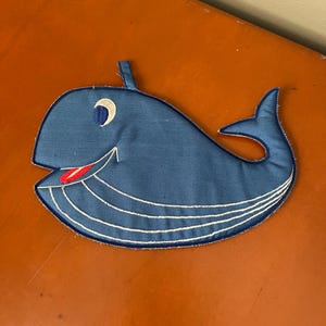 May include: A blue whale-shaped applique with white stitching details. The whale has a smiling face with a red tongue, a white crescent eye, and a small fin. The applique is likely made of fabric and is designed for sewing onto clothing or other items.