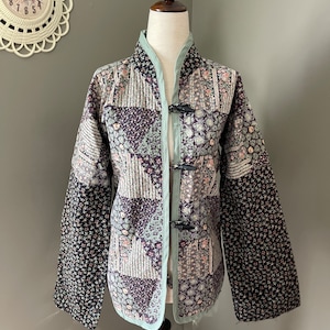 May include: A quilted jacket with a patchwork design, displayed on a mannequin. The jacket features a variety of floral and geometric patterns in shades of black, purple, and cream. It has a mandarin collar and toggle closures. The trim is a light green color.