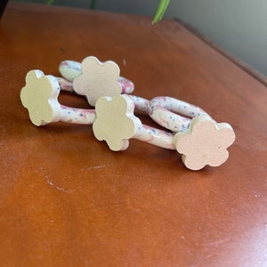 May include: Three decorative ceramic flower-shaped drawer pulls. The pulls have a white base with a floral pattern of pink and blue dots. The flower-shaped ends are a light beige color. The pulls are resting on a brown surface.