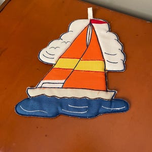 May include: A fabric sailboat applique with an orange and yellow sail, white clouds, and blue waves. The sailboat has a white hull and a red accent at the top of the sail. A loop is attached at the top for hanging.