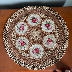 May include: A round, woven placemat with a decorative border. The placemat features six circular embroidered coasters with red and pink floral designs. A central floral design is also present.