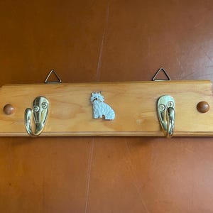 May include: A wooden coat rack with two gold-colored hooks and a white fox figurine in the center. The rack has a light wood finish with two small wooden knobs and two metal triangle hangers.