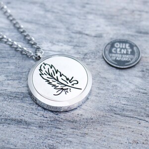 May include: A silver-colored pendant necklace with a circular pendant featuring a black feather design. The necklace includes a chain and is shown next to a United States one-cent coin.