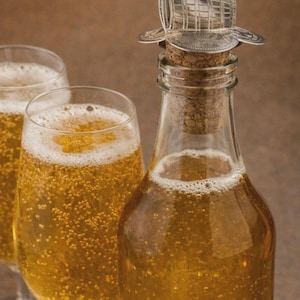Peut inclure: Une bouteille en verre clair et deux verres remplis d'un liquide doré et pétillant, probablement du cidre. La bouteille est coiffée d'un bouchon de liège et d'un bouchon décoratif en métal avec le mot "CIDER".