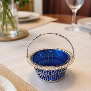 Puede incluir: Una taza de cerámica blanca con un asa negra y el texto "But First, Coffee" en negrita negra. La taza está sobre un fondo borroso. La taza mide aproximadamente 10 cm de altura.