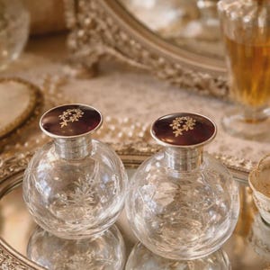 May include: Two clear, round perfume bottles with silver collars and dark red stoppers. The stoppers have a gold floral design. The bottles sit on a mirrored tray, reflecting their shape. The background includes a mirror and other decorative items.
