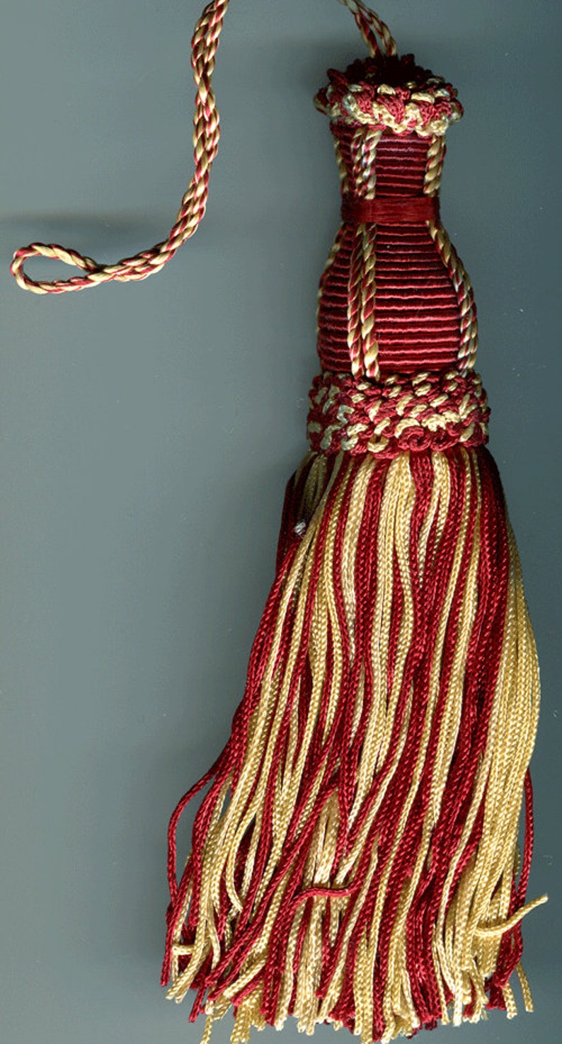 May include: A red and gold tassel with a long cord. The tassel is made of braided cords and has a fringe of long strands.