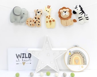 May include: A felt animal banner with an elephant, cheetah, giraffe, lion, and zebra. A white star-shaped light and a framed rainbow with a heart are also present. The text "WILD at heart" is on a card, with felt balls below.