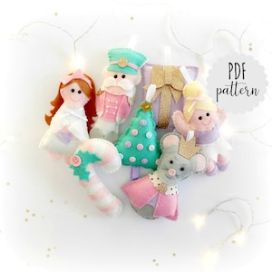 May include: A selection of handmade felt Christmas decorations, featuring a nutcracker, a ballerina, a candy cane, a Christmas tree, a gift, and a mouse. The ornaments are mainly pink, white, and green, with gold detailing. The text "PDF pattern" is visible.