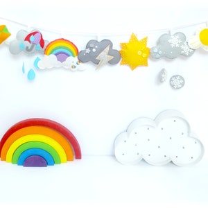 K&ouml;nnte beinhalten: Eine Filzgirlande mit einem Regenbogen, Wolken, einer Sonne, einem Blitz und Regentropfen. Au&szlig;erdem gibt es einen Holzregenbogen und eine wei&szlig;e Wolke mit silbernen Glitzer.