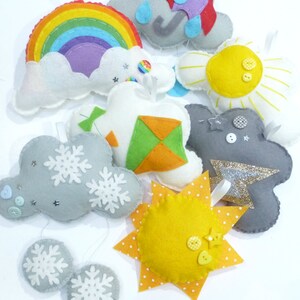 K&ouml;nnte beinhalten: Ein Set aus Filz-Anh&auml;ngern, die verschiedene Wetterbedingungen darstellen, darunter ein Regenbogen, eine Wolke mit Regen, eine Sonne, eine Wolke mit Schneeflocken und ein Drachen. Die Anh&auml;nger sind mit Kn&ouml;pfen und Glitzer verziert.