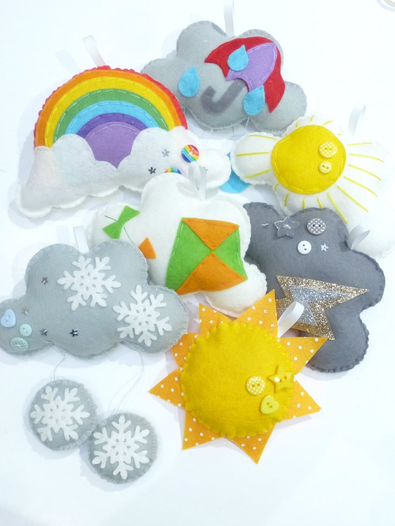 K&ouml;nnte beinhalten: Ein Set aus Filz-Anh&auml;ngern, die verschiedene Wetterbedingungen darstellen, darunter ein Regenbogen, eine Wolke mit Regen, eine Sonne, eine Wolke mit Schneeflocken und ein Drachen. Die Anh&auml;nger sind mit Kn&ouml;pfen und Glitzer verziert.